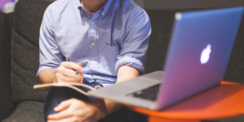 man sitting in front of laptop writing into notebook in lap