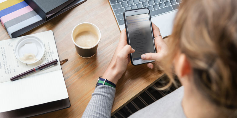 over the shoulder of woman with smartphone in hand at desk with coffee, notebooks, and laptop