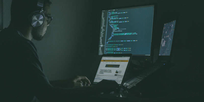 man with headphones in dark room using two laptops and an external monitor for web development