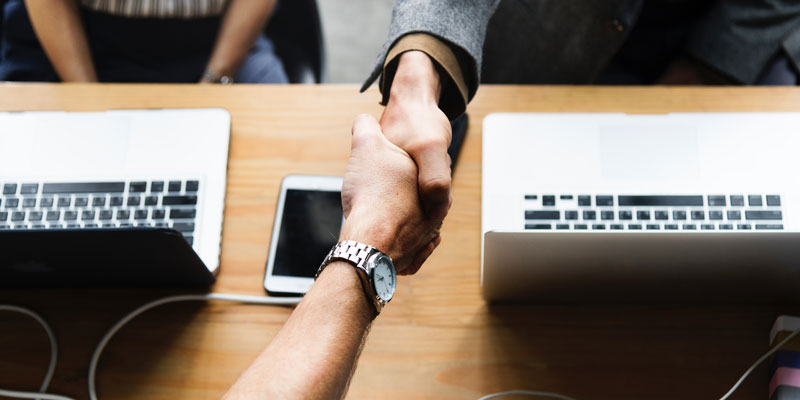 overhead view of handshake across wooden table of 2 laptops and 1 cell phone