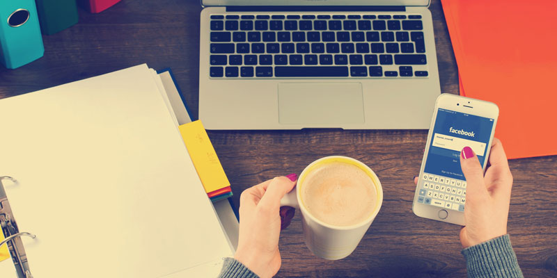 woman's viewpoint sitting in front of laptop and folders, holding cup of coffee and smartphone using facebook