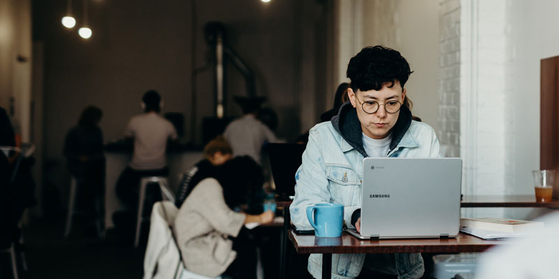 woman wearing jean jacket with a mug and working on computer in coffee shop