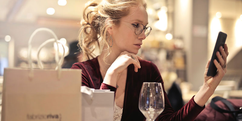 woman on phone sitting at table with shopping bags and empty glass of wine